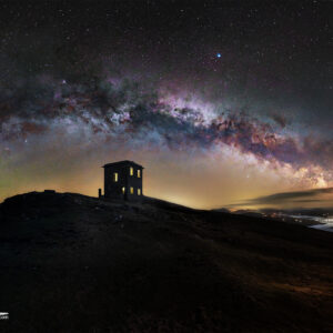 Milky Way galaxy arching over Bray Head, Ireland, with a solitary house glowing on a hill under a star-filled night sky.